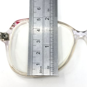 Pu&ograve; includere: Occhiali in plastica trasparente con un accento rosa sulla parte superiore della montatura. Gli occhiali sono misurati con un righello, mostrando che la larghezza della montatura &egrave; di circa 5 cm.