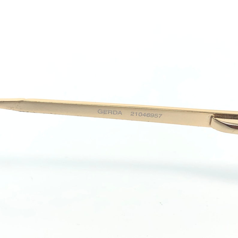 May include: Close-up of a gold-colored metal object with the text "GERDA 21046957" etched on its surface. The object has a sleek, modern design and appears to be a part of a larger item, possibly eyewear or jewelry.