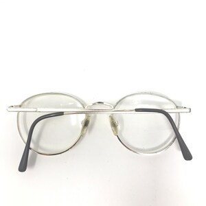 Peut inclure: Lunettes &agrave; monture argent&eacute;e avec verres transparents. Les lunettes ont une forme arrondie et des branches noires. Les plaquettes nasales sont transparentes. Les lunettes sont pos&eacute;es sur un fond blanc.