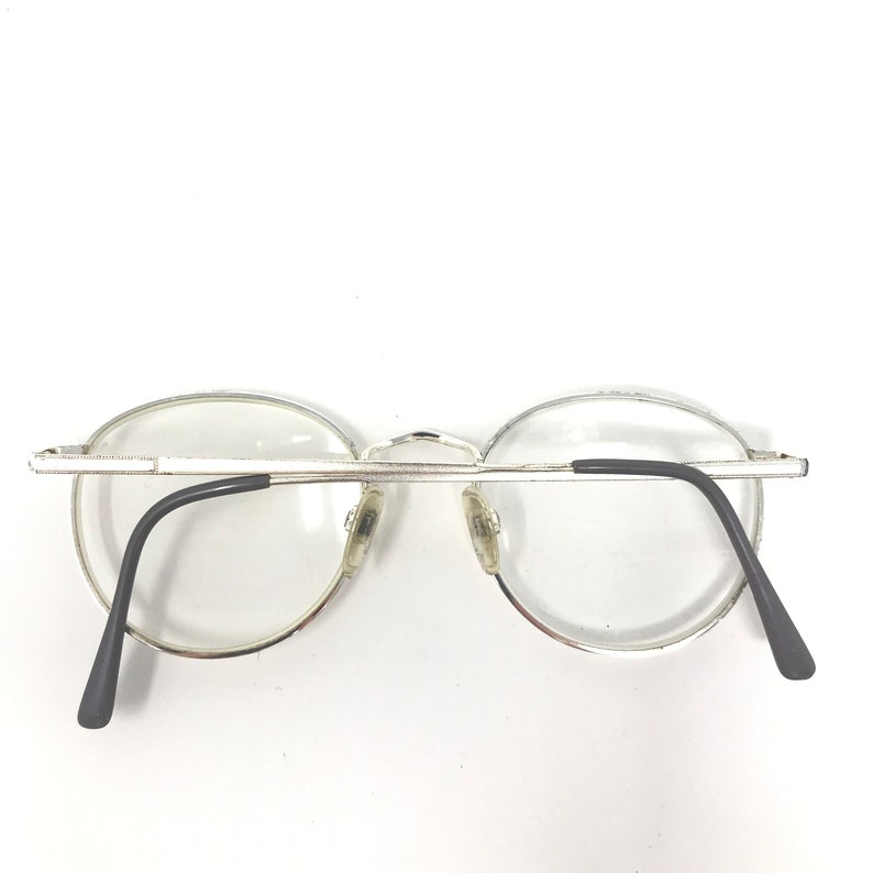 Peut inclure: Lunettes &agrave; monture argent&eacute;e avec verres transparents. Les lunettes ont une forme arrondie et des branches noires. Les plaquettes nasales sont transparentes. Les lunettes sont pos&eacute;es sur un fond blanc.