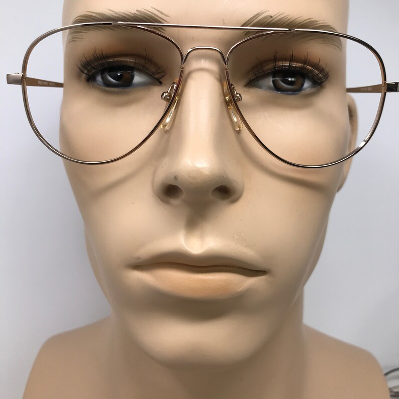 May include: Gold-tone aviator eyeglasses with clear lenses. The glasses have a double bridge and a classic teardrop shape. The nose pads are a light beige color. The glasses are displayed on a mannequin.