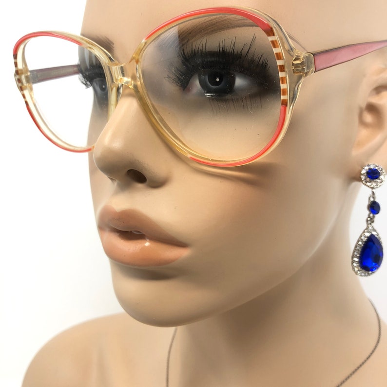 Puede incluir: Gafas vintage con montura transparente, acentuadas con un borde rojo. Las gafas tienen un estilo retro, con una forma grande y redondeada. La imagen tambi&eacute;n muestra un par de pendientes colgantes azules y plateados.