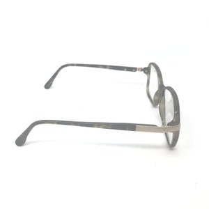 Puede incluir: Un par de gafas con montura moteada en verde oscuro y gris. Las patillas son de color verde oscuro con un adorno plateado cerca de las bisagras. Las lentes son transparentes.