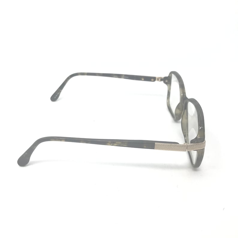 Puede incluir: Un par de gafas con montura moteada en verde oscuro y gris. Las patillas son de color verde oscuro con un adorno plateado cerca de las bisagras. Las lentes son transparentes.