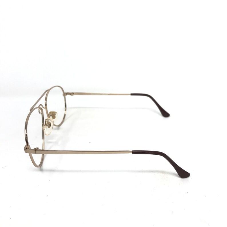 May include: A pair of silver-toned aviator eyeglasses with clear lenses. The frames have a double bridge and brown temple tips. The glasses are set against a white background.