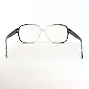 K&ouml;nnte beinhalten: Eine Vintage-Brille mit einem Farbverlauf-Rahmen, der von Schwarz zu Klar &uuml;bergeht. Die quadratischen Gl&auml;ser sind von einem dicken Rahmen umgeben, und die B&uuml;gel sind dunkelgrau. Die Brille ist vor einem wei&szlig;en Hintergrund platziert.