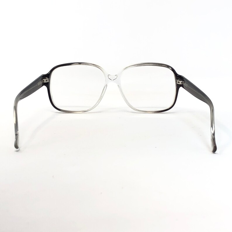 K&ouml;nnte beinhalten: Eine Vintage-Brille mit einem Farbverlauf-Rahmen, der von Schwarz zu Klar &uuml;bergeht. Die quadratischen Gl&auml;ser sind von einem dicken Rahmen umgeben, und die B&uuml;gel sind dunkelgrau. Die Brille ist vor einem wei&szlig;en Hintergrund platziert.