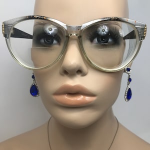 May include: A pair of vintage cat-eye glasses with a silver and black frame and clear lenses. The glasses are on a mannequin's face. The mannequin is wearing blue teardrop earrings.