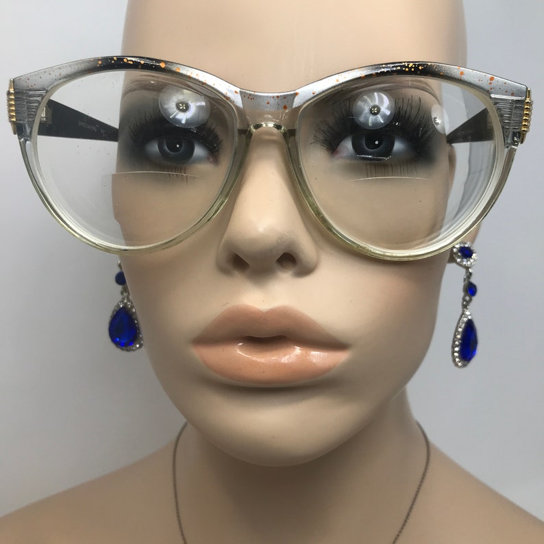 May include: A pair of vintage cat-eye glasses with a silver and black frame and clear lenses. The glasses are on a mannequin's face. The mannequin is wearing blue teardrop earrings.