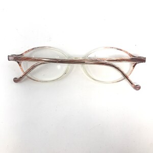 May include: Oval-shaped spectacles with clear lenses and a brown and silver patterned frame. The temples curve gently, ending in rounded tips. The spectacles are set against a white background.