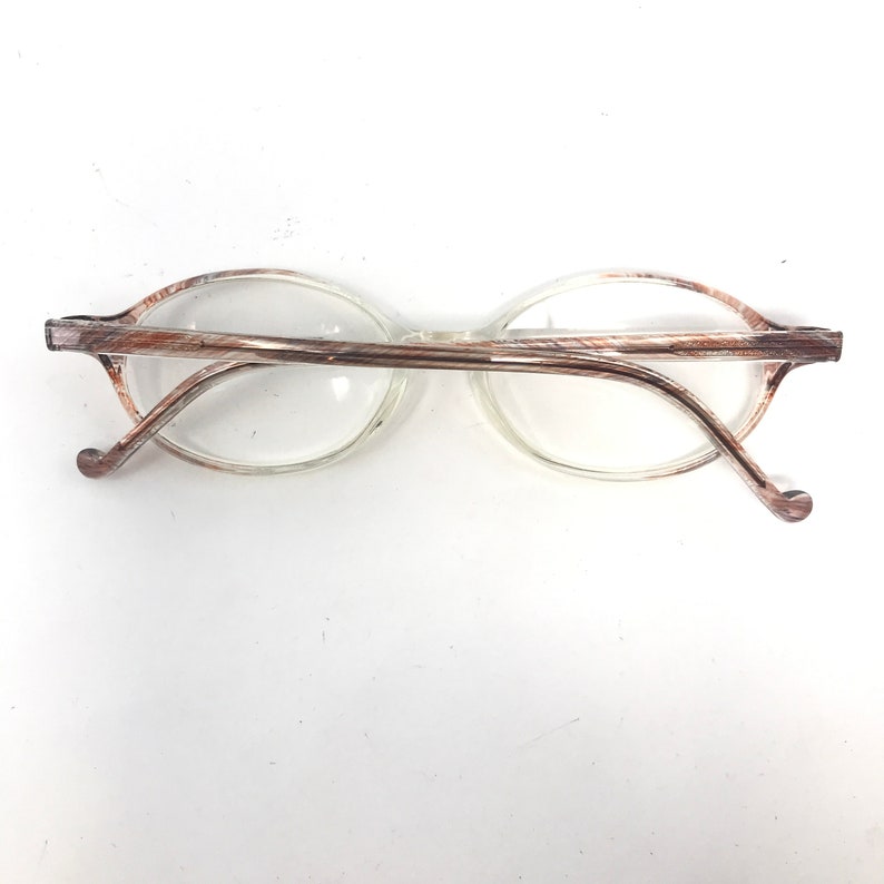 May include: Oval-shaped spectacles with clear lenses and a brown and silver patterned frame. The temples curve gently, ending in rounded tips. The spectacles are set against a white background.