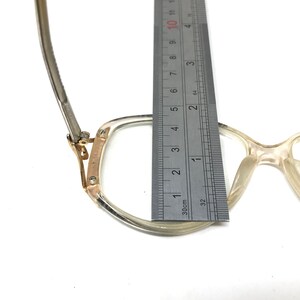 Puede incluir: Primer plano de un par de gafas de pl&aacute;stico transparente con una bisagra de metal dorado. Las gafas se miden con una regla, mostrando que el ancho del marco es de aproximadamente 4,5 cm.