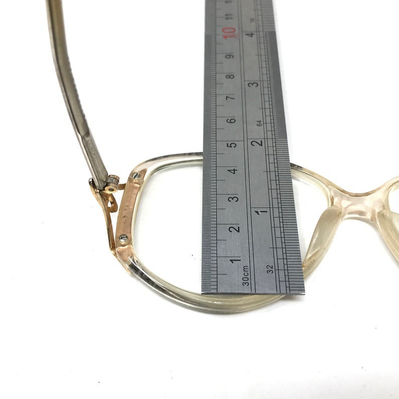 Puede incluir: Primer plano de un par de gafas de pl&aacute;stico transparente con una bisagra de metal dorado. Las gafas se miden con una regla, mostrando que el ancho del marco es de aproximadamente 4,5 cm.