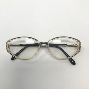 Puede incluir: Gafas de forma ovalada con lentes transparentes y montura marr&oacute;n claro. Las patillas son negras con detalles dorados y el texto "CHARLENE" es visible en la patilla izquierda. Las gafas est&aacute;n sobre un fondo blanco.