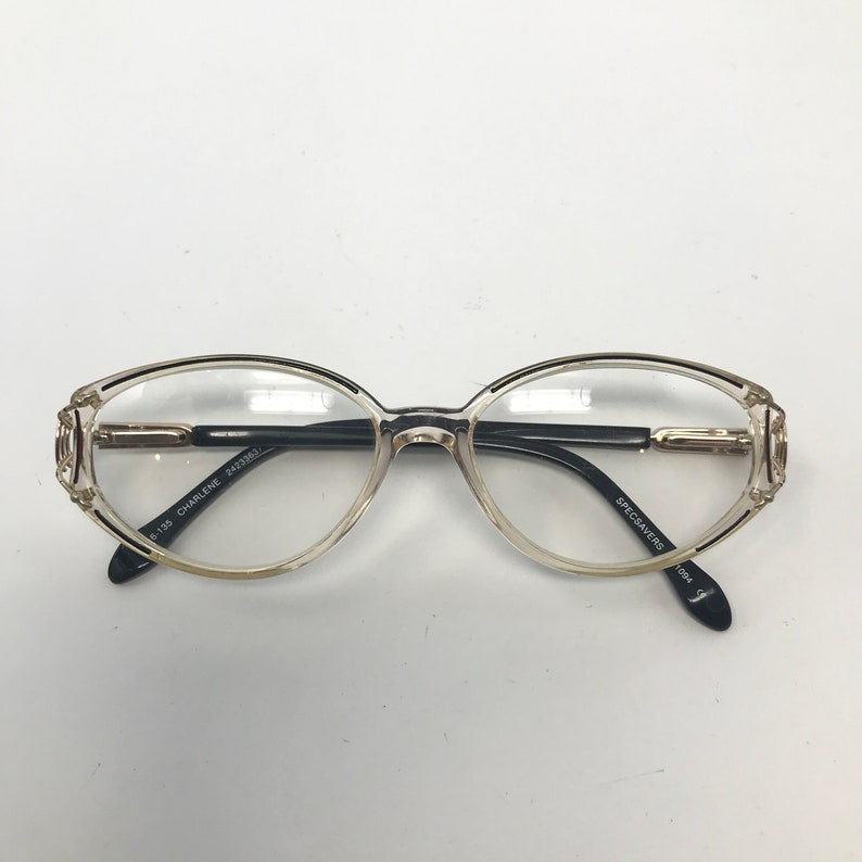Puede incluir: Gafas de forma ovalada con lentes transparentes y montura marr&oacute;n claro. Las patillas son negras con detalles dorados y el texto "CHARLENE" es visible en la patilla izquierda. Las gafas est&aacute;n sobre un fondo blanco.