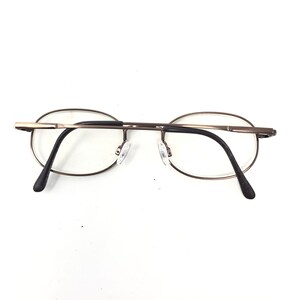 Puede incluir: Gafas de forma ovalada con una fina montura de metal color bronce. Las lentes son transparentes y las patillas son negras. Las almohadillas nasales son transparentes. Las gafas est&aacute;n sobre un fondo blanco.