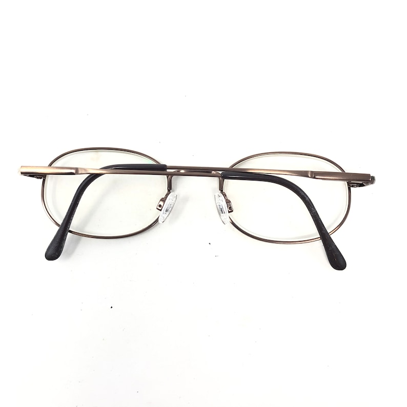Puede incluir: Gafas de forma ovalada con una fina montura de metal color bronce. Las lentes son transparentes y las patillas son negras. Las almohadillas nasales son transparentes. Las gafas est&aacute;n sobre un fondo blanco.