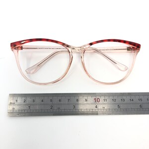 May include: Clear eyeglasses with a light pink frame and a tortoiseshell pattern on the upper rims. A metal ruler is placed below the glasses, with measurements in inches and centimeters.