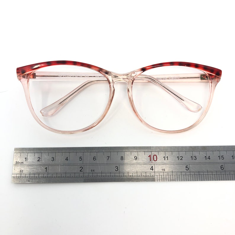 May include: Clear eyeglasses with a light pink frame and a tortoiseshell pattern on the upper rims. A metal ruler is placed below the glasses, with measurements in inches and centimeters.