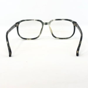 May include: Eyeglasses with a gray and white speckled pattern. The frames are square-shaped with clear lenses. The temples curve outward, and the glasses are set against a white background.
