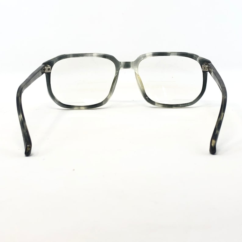 May include: Eyeglasses with a gray and white speckled pattern. The frames are square-shaped with clear lenses. The temples curve outward, and the glasses are set against a white background.