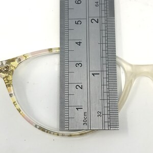 May include: Close-up of eyeglasses with a clear lens and a patterned frame. A ruler is placed over the lens, measuring approximately 2 inches. The frame is a mix of light colors, including yellow, pink, and white.