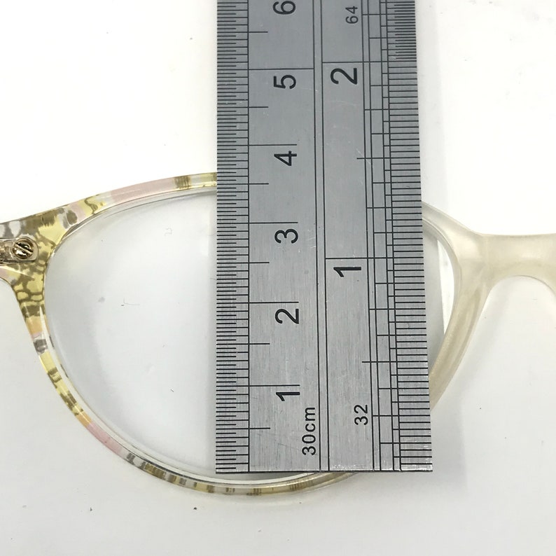May include: Close-up of eyeglasses with a clear lens and a patterned frame. A ruler is placed over the lens, measuring approximately 2 inches. The frame is a mix of light colors, including yellow, pink, and white.