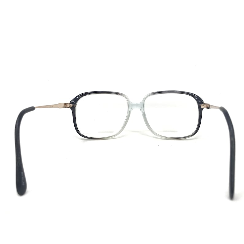 May include: Square-framed spectacles with a gradient black to clear frame. The temples are silver-toned metal, and the earpieces are black. The glasses are set against a white background.