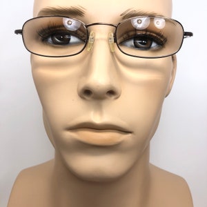 Puede incluir: Gafas rectangulares marrones oscuras con lentes transparentes, presentadas en una cabeza de maniqu&iacute;. Las gafas tienen una montura de metal fina y un peque&ntilde;o puente nasal. El estilo es cl&aacute;sico y discreto. El fondo es blanco.