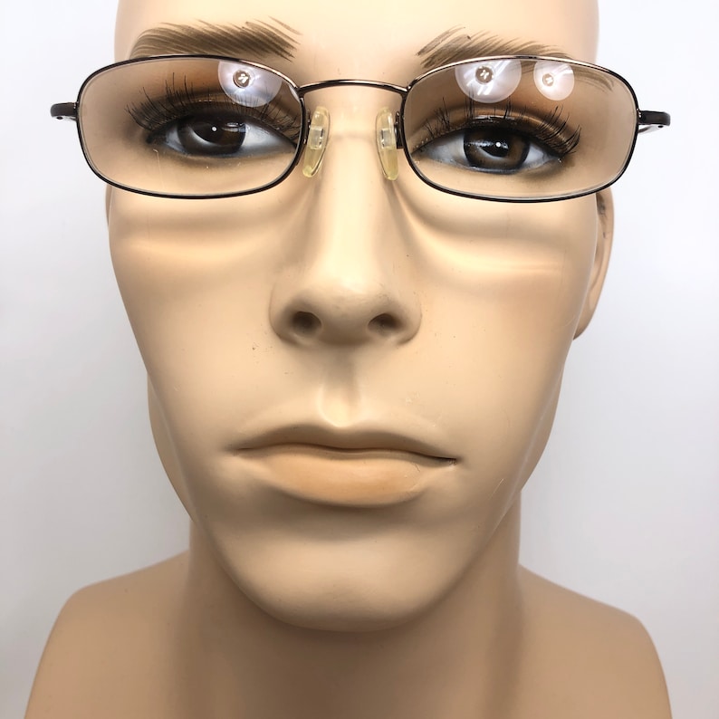 Puede incluir: Gafas rectangulares marrones oscuras con lentes transparentes, presentadas en una cabeza de maniqu&iacute;. Las gafas tienen una montura de metal fina y un peque&ntilde;o puente nasal. El estilo es cl&aacute;sico y discreto. El fondo es blanco.