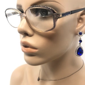 May include: Vintage eyeglasses with a light brown frame and clear lenses. The glasses are paired with blue and silver drop earrings and a delicate necklace. The earrings feature a round top, three blue stones, and a teardrop-shaped bottom stone.
