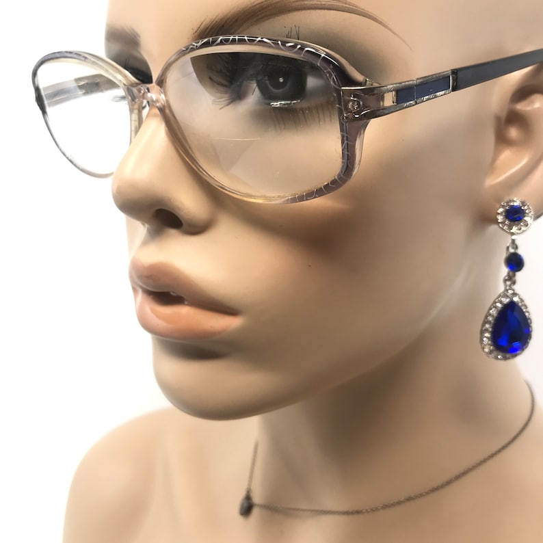 May include: Vintage eyeglasses with a light brown frame and clear lenses. The glasses are paired with blue and silver drop earrings and a delicate necklace. The earrings feature a round top, three blue stones, and a teardrop-shaped bottom stone.