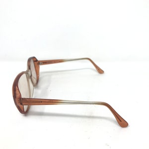 May include: A pair of vintage eyeglasses with a light brown frame and clear lenses. The arms of the glasses are a gradient, transitioning from brown to clear. The glasses are set against a white background.
