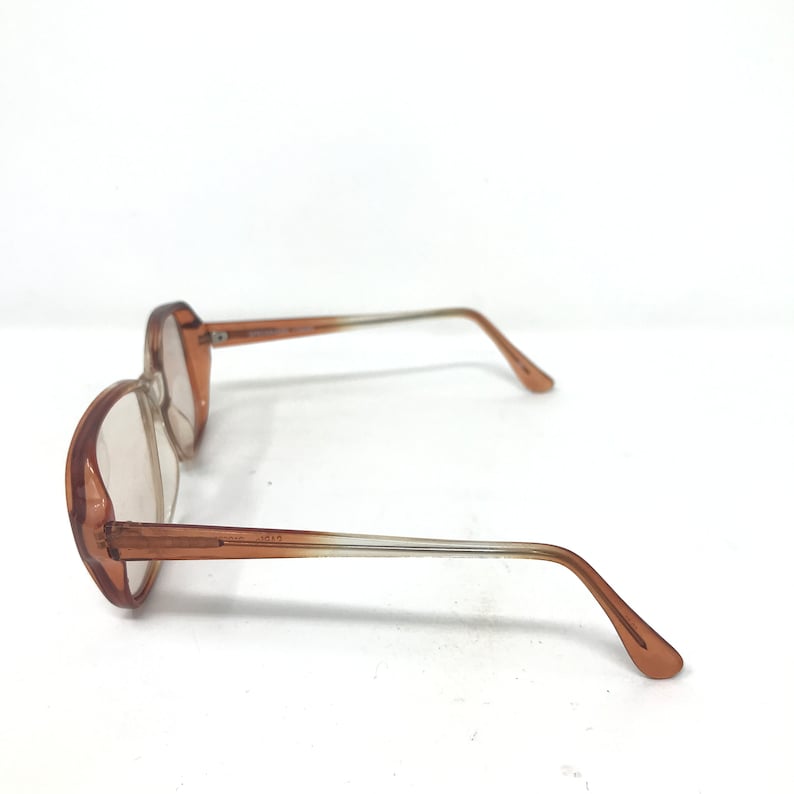 May include: A pair of vintage eyeglasses with a light brown frame and clear lenses. The arms of the glasses are a gradient, transitioning from brown to clear. The glasses are set against a white background.