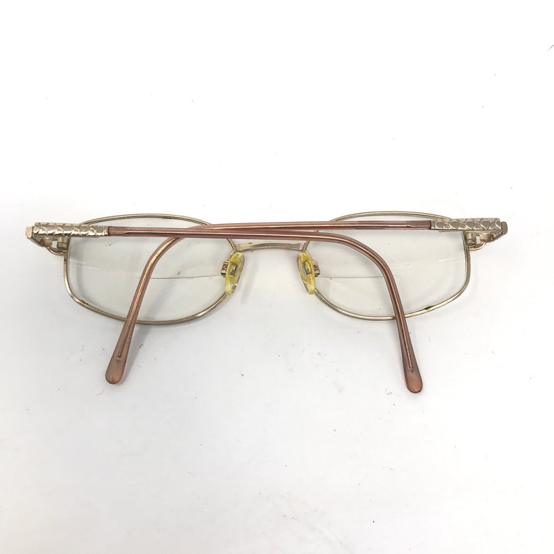 May include: Eyeglasses with a gold-coloured frame and clear lenses. The temples and bridge are a light brown colour. The frame has decorative detailing on the temples.