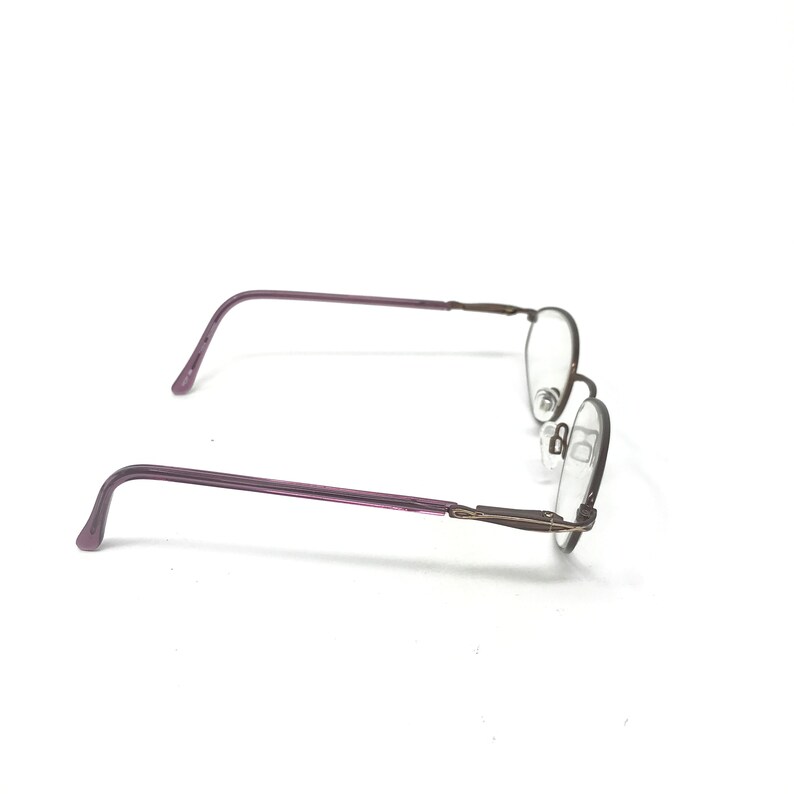 Puede incluir: Un par de gafas con una montura fina y ovalada. Las patillas son de color p&uacute;rpura apagado y la montura es de color oscuro y met&aacute;lico. Las lentes son transparentes y se ven las almohadillas nasales.