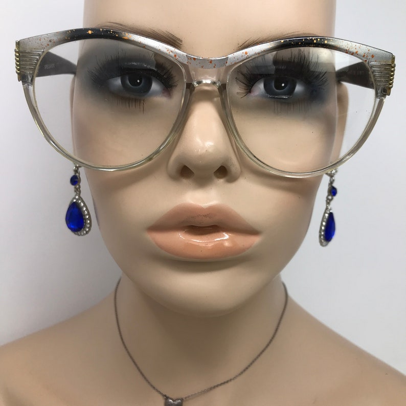May include: Clear-framed eyeglasses with a cat-eye shape, featuring gold accents and speckled detailing. The model is also wearing blue teardrop earrings with silver accents. The overall aesthetic is vintage and glamorous.