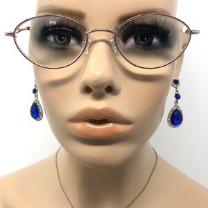 Puede incluir: Un busto de maniqu&iacute; con gafas ovaladas de oro rosa y pendientes colgantes de l&aacute;grima azules. Los pendientes tienen un engaste plateado con cristales transparentes alrededor de la piedra azul. El maniqu&iacute; tiene largas pesta&ntilde;as y un labial neutro.