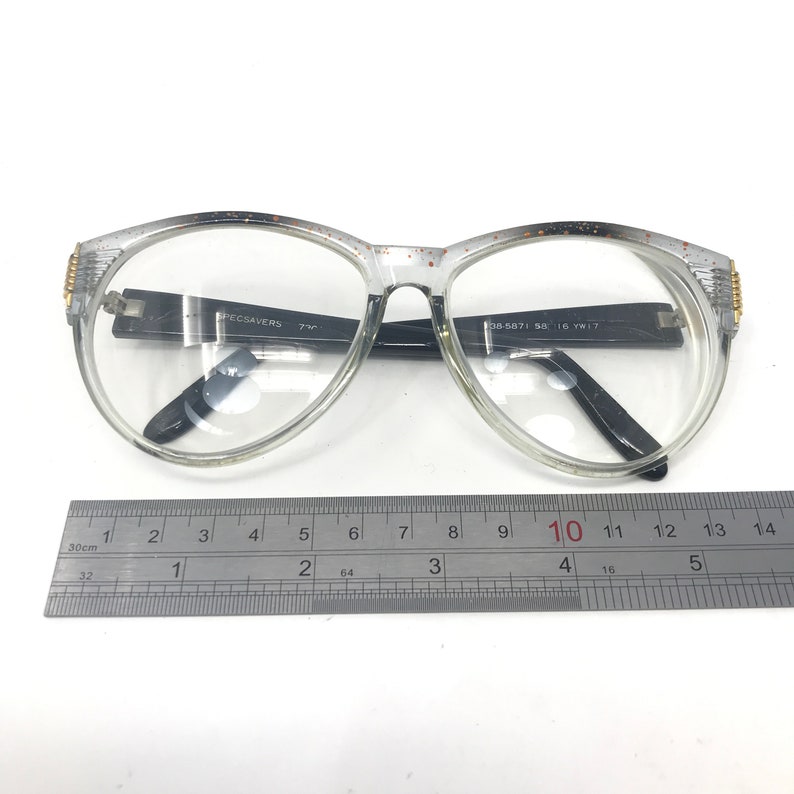 May include: Clear plastic eyeglasses with black arms and gold accents. The arms are labeled "SPECSAVERS 73C" and "08-5871 58 16 YW17". The eyeglasses are measured on a ruler, showing they are approximately 10 centimeters wide.