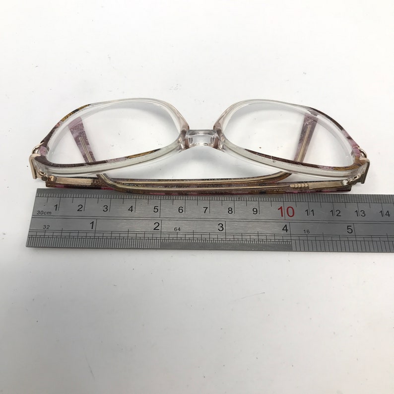 May include: Clear-framed eyeglasses with a rectangular shape, featuring a decorative pattern on the temples. The glasses are approximately 5.5 inches wide, measured with a ruler. The lenses are clear, and the frame is a mix of clear and patterned materials.