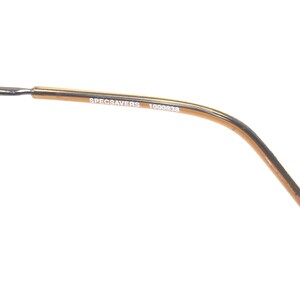 Puede incluir: Primer plano de una patilla de gafas dorada. El texto "SPECSAVERS 1000838" est&aacute; impreso en una fuente peque&ntilde;a y sin serifa en la patilla. La montura se curva suavemente, con una fina patilla plateada que se extiende desde el extremo.