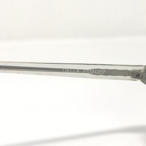 May include: Close-up of a clear eyeglass arm. The text "DELLA 21024207" is printed on the arm. The arm is made of a transparent material and is set against a white background. The image is well-lit and focused.