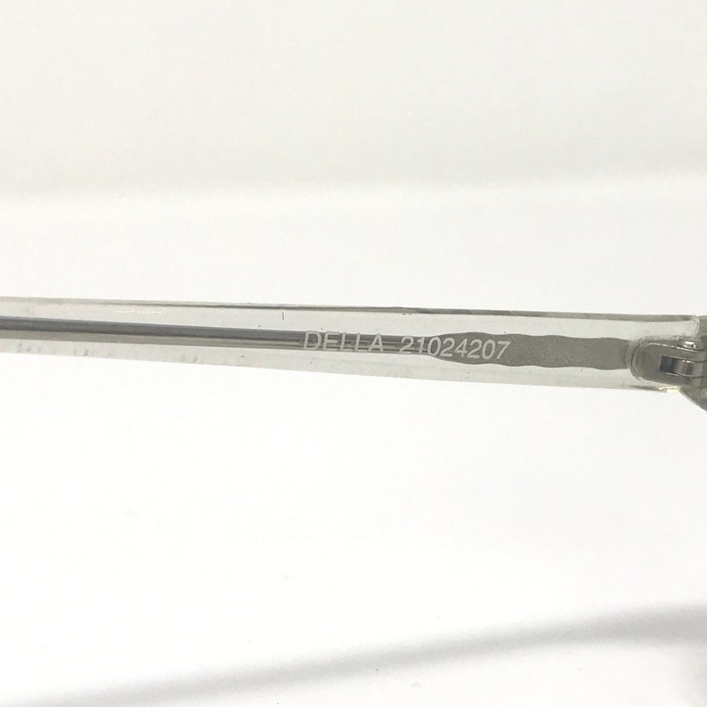 May include: Close-up of a clear eyeglass arm. The text "DELLA 21024207" is printed on the arm. The arm is made of a transparent material and is set against a white background. The image is well-lit and focused.