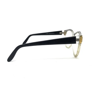 May include: A pair of eyeglasses with a clear frame and black temples. The temples are connected to the frame with a gold-colored accent. The lenses are clear, and the glasses are set against a white background.