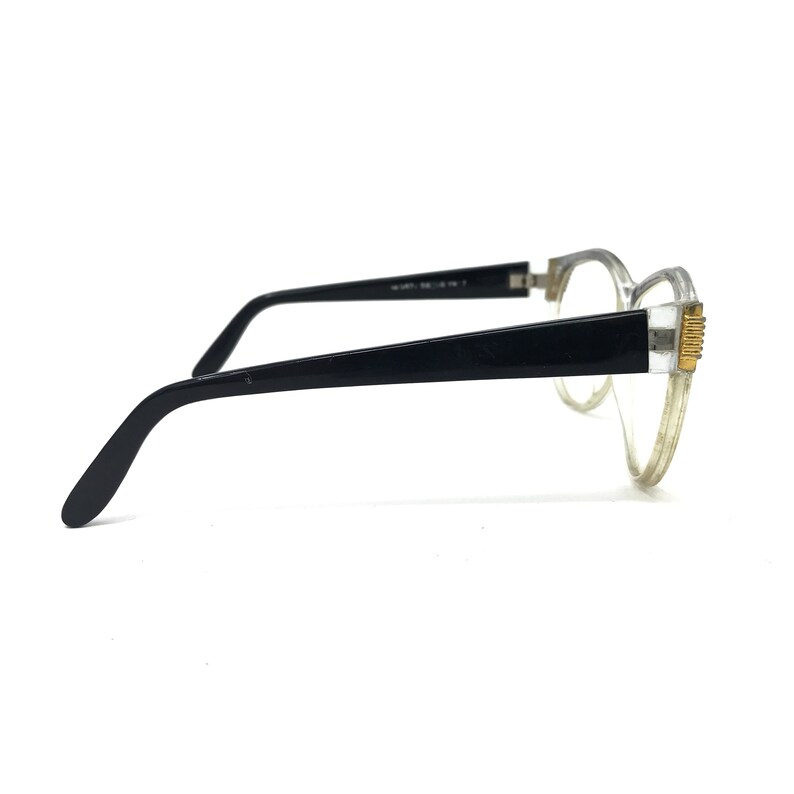 May include: A pair of eyeglasses with a clear frame and black temples. The temples are connected to the frame with a gold-colored accent. The lenses are clear, and the glasses are set against a white background.