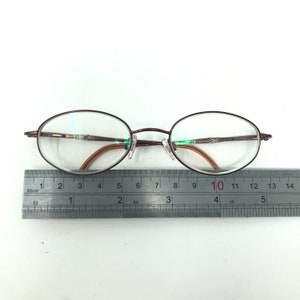 May include: Oval-shaped eyeglasses with a dark brown metal frame. The lenses are clear, and the nose pads are transparent. A ruler is placed below the glasses, showing measurements in inches.