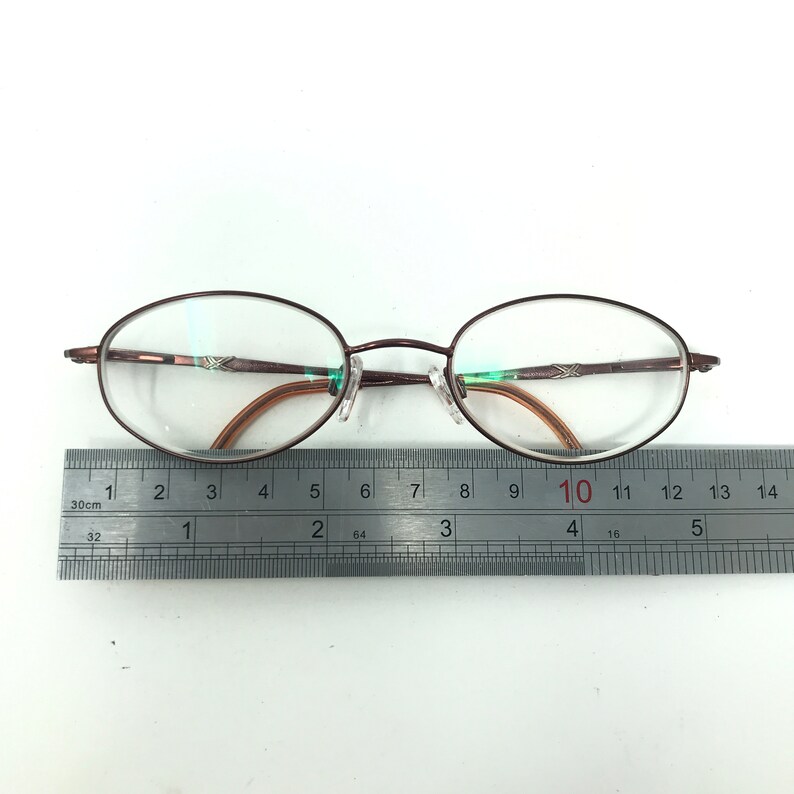 May include: Oval-shaped eyeglasses with a dark brown metal frame. The lenses are clear, and the nose pads are transparent. A ruler is placed below the glasses, showing measurements in inches.