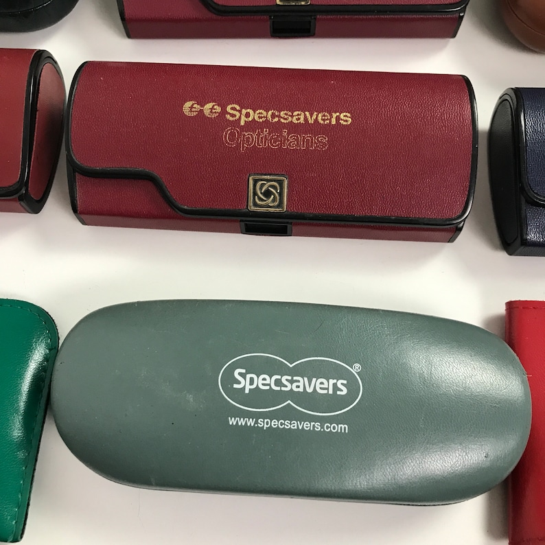 May include: An assortment of eyeglass cases in various colors, including red, green, and gray. The cases feature the "Specsavers" logo and branding. Some cases are rectangular with a snap closure, while others are oval-shaped.