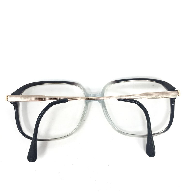 May include: Eyeglasses with a black and clear frame and black temples. The glasses have a rectangular shape with a silver-coloured metal bridge and accents. The lenses are clear.