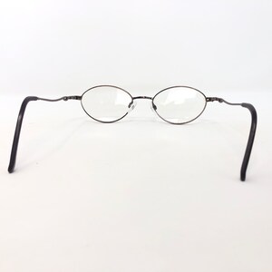 Peut inclure: Lunettes de vue ovales avec une monture en m&eacute;tal fonc&eacute; et des verres transparents. Les branches sont fines et incurv&eacute;es au niveau des branches. Les lunettes sont pos&eacute;es sur un fond blanc.